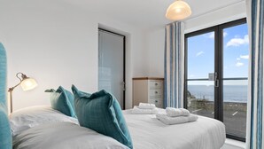 A bedroom with a large bed, a nightstand, a dresser, and a view of the sea through a large window. - Apartment 8 Waterstone House - Luxury Apartment Sea Views Pet Friendly (Tenby)