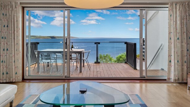 A modern living room with a glass table, a sofa, and a view of the sea through large sliding doors. - Cystanog Fach - Spectacular Sea Views Balcony and Terrace Parking (Saundersfoot)