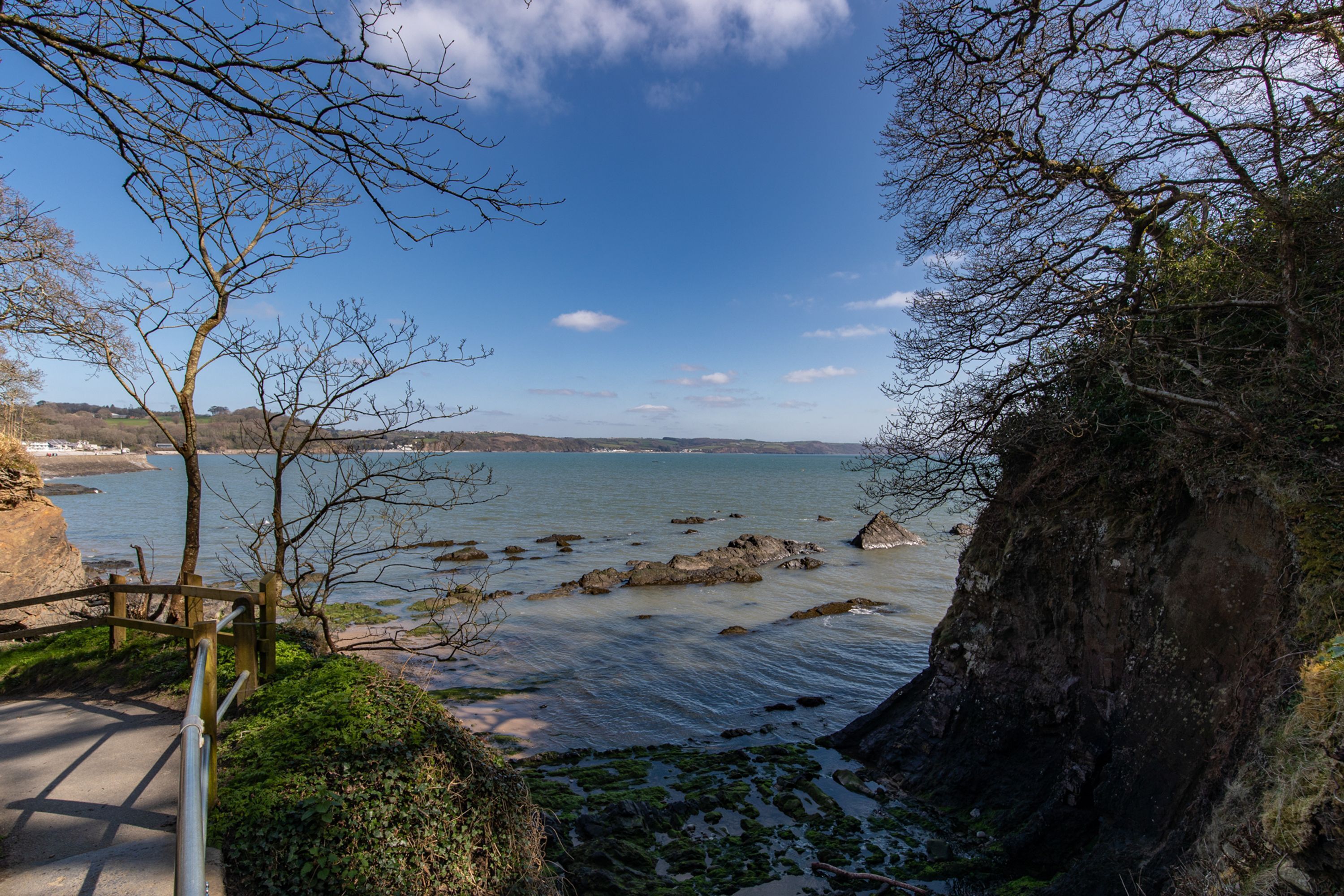 A coastal landscape with a rocky shoreline, a wooden pathway, and a view of the sea and distant hills.