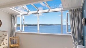 A bedroom with a large bay window overlooking a body of water, a wooden chair, and a bed with pillows. - Cystanog Fach - Spectacular Sea Views Balcony and Terrace Parking (Saundersfoot)