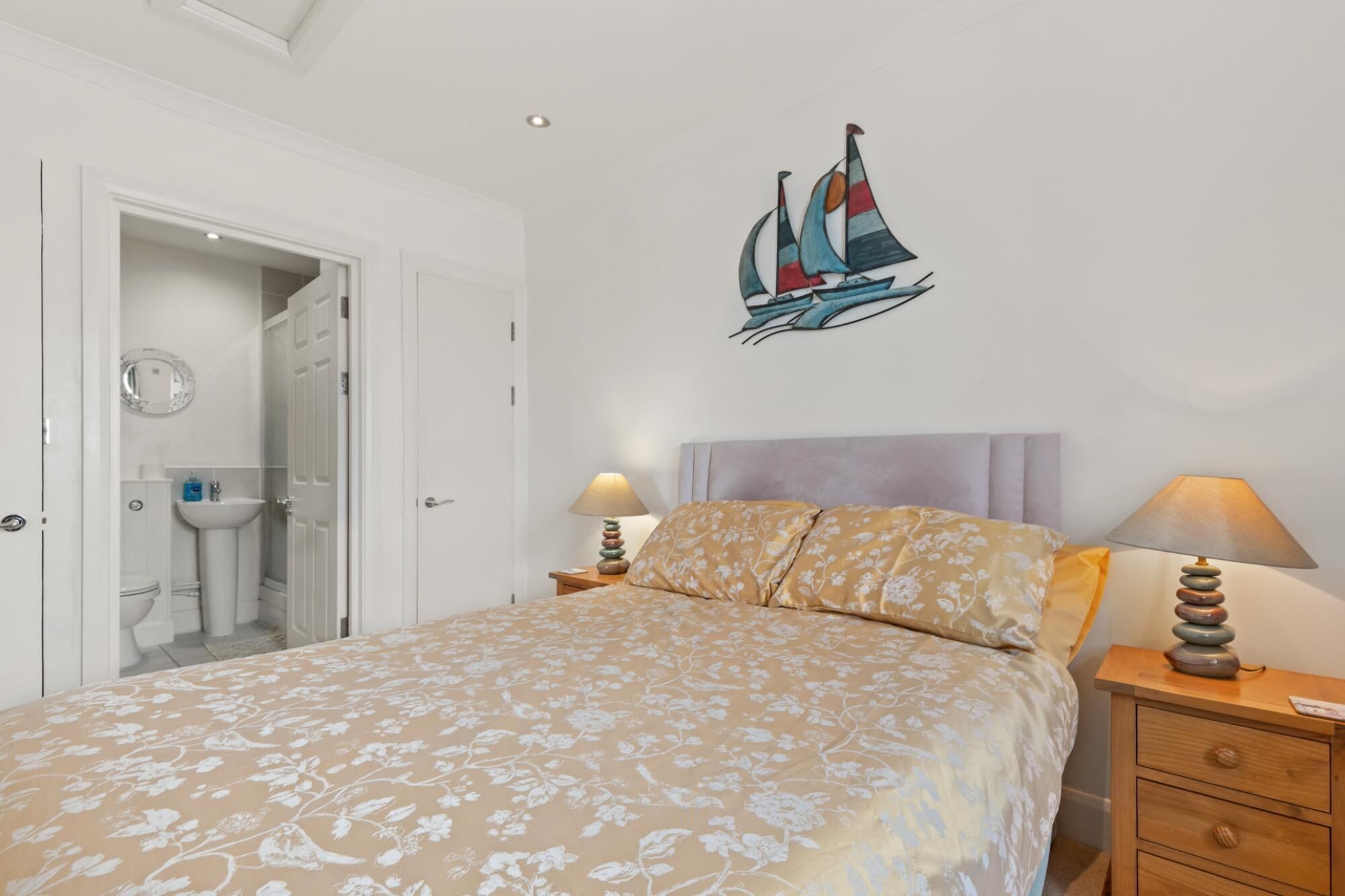 A bedroom with a bed, two bedside tables with lamps, a wall decoration of sailboats, a bathroom with a mirror and sink, and a door leading to another room.