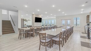 A spacious dining area with a long table and multiple chairs, a staircase, and a kitchen area with a large window. - THE OCEANFRONT GRAND RESORT: 17 BR Luxury with Unmatched Views+ALL THE AMENITIES (North Myrtle Beach)