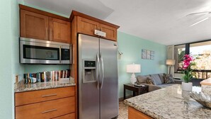 A kitchen with wooden cabinets, a stainless steel refrigerator, a microwave, and a granite countertop. - Beach theme, private Lanai, pools and tennis courts (Kihei)