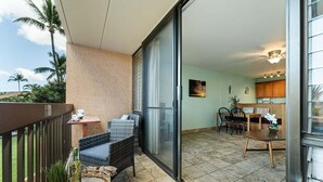 A balcony with wicker furniture, a dining area with wooden chairs, and a kitchen visible through a glass door. - Beach theme, private Lanai, pools and tennis courts (Kihei)