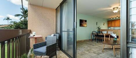 A balcony with wicker furniture, a dining area with wooden chairs, and a kitchen visible through a glass door.