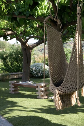 A hammock suspended from a tree in a garden setting with a bench and a stone wall in the background.