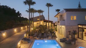 A poolside area with lounge chairs, a swimming pool, and a building with a stone facade and palm trees. - Kipo paradiso villa, pool,spa,massage,yoga,gym (Kyrianna)
