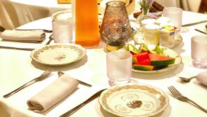 A breakfast setting with a table set for two, including plates, cutlery, a glass of orange juice, a bottle of water, and a fruit bowl.