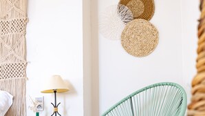 A room with a white wall, a macrame wall hanging, a green woven chair, a small white nightstand with a lamp, and a woven wall art piece.