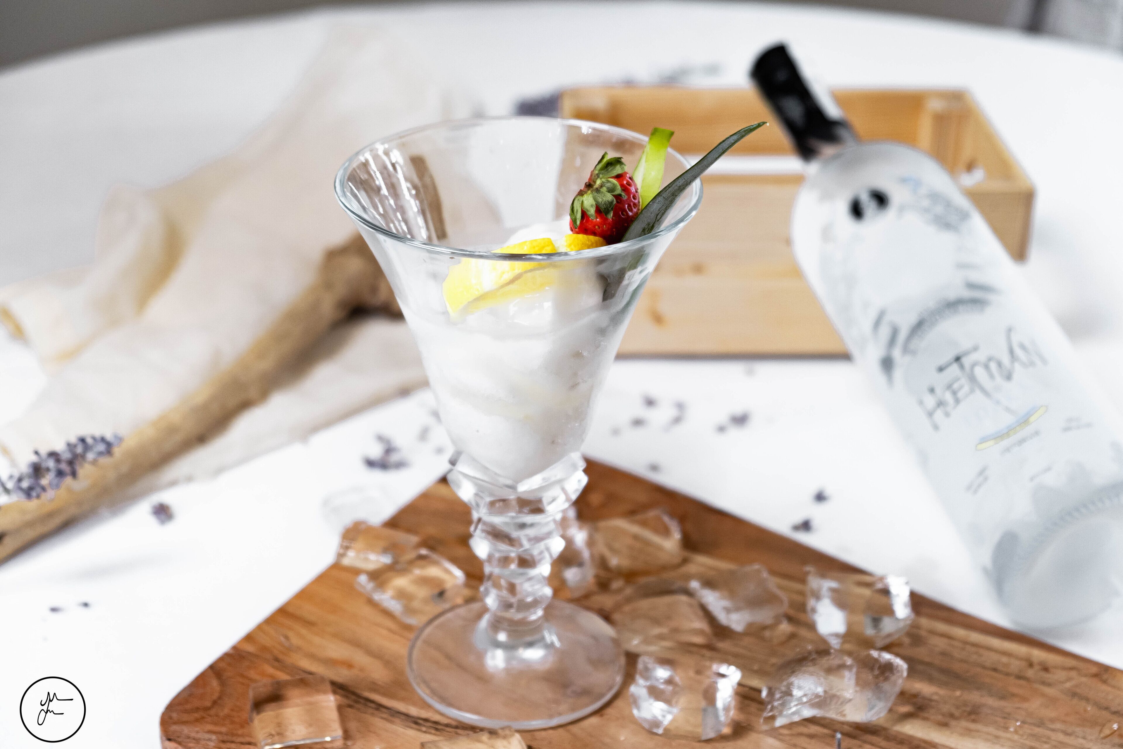 Ett martiniglas med en citronskiva och en jordgubbe som garnering, placerat på en träbricka. I bakgrunden syns isbitar och en flaska Hennessy-cognac.