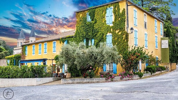 A yellow building with blue shutters, surrounded by greenery and a clear sky.