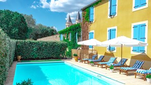 A swimming pool with lounge chairs and umbrellas, surrounded by greenery and a building with blue shutters.