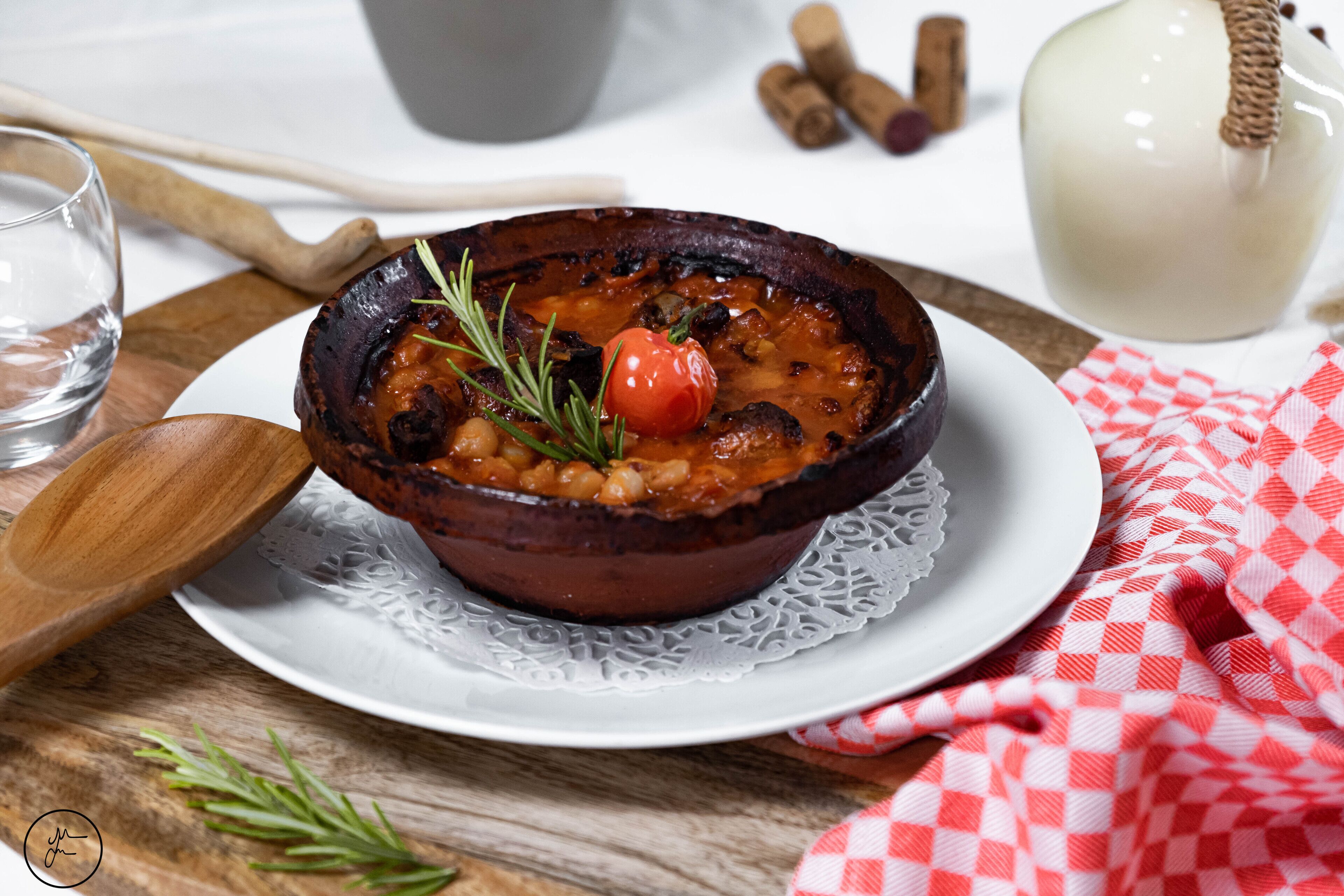 A bowl of food on a white plate with a lace doily, wooden spoon, and a red and white checkered cloth.