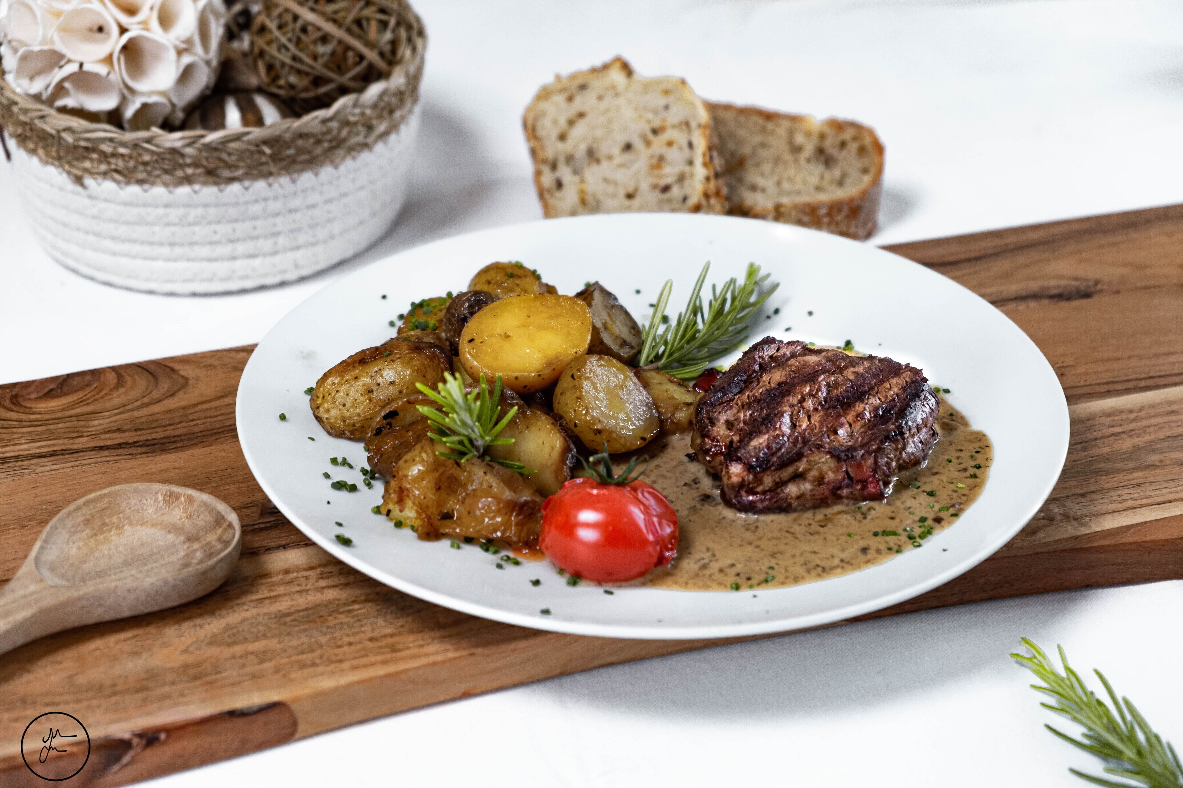 A plate of food with steak, potatoes, and a cherry tomato, accompanied by slices of bread on a wooden table.