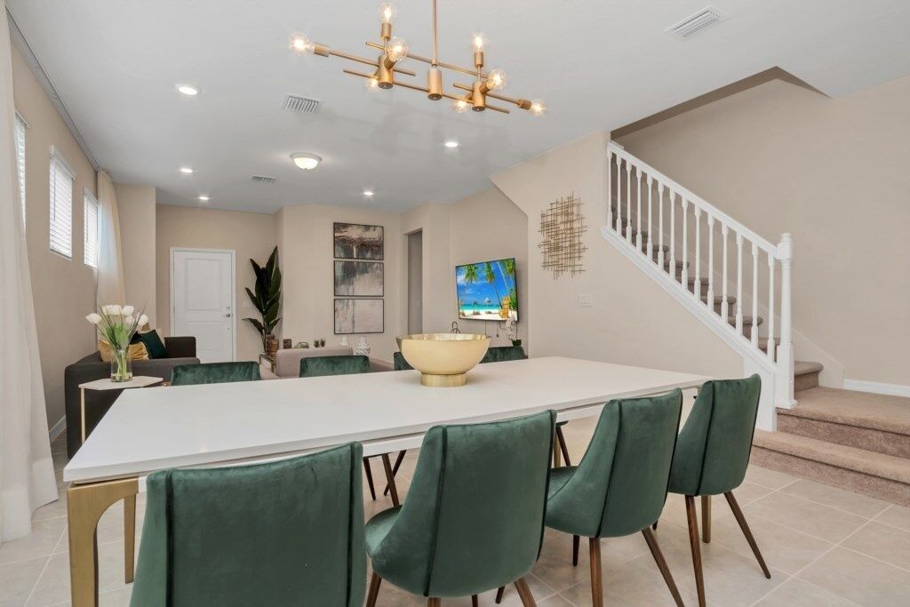 A dining area with a long table, green upholstered chairs, a central island with a bowl, and a staircase in the background.