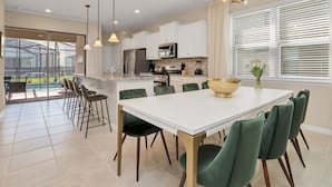 A modern kitchen with white cabinets, a large island, and a dining area with green chairs. - Best Resort Near Disney Private Pool & Spa Home! 5 Bedroom Villa by RedAwning (Davenport)
