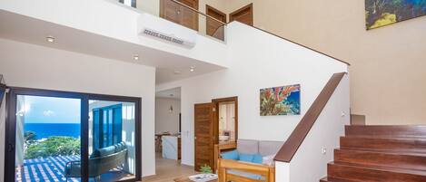 A spacious living area with a wooden staircase, a balcony with a view of the ocean, and a dining area.