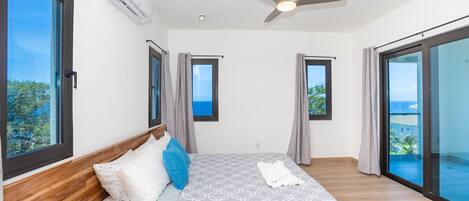 Une chambre dotée d’un grand lit, d’un ventilateur de plafond et offrant une vue sur l’océan par le biais d’une porte-fenêtre coulissante.