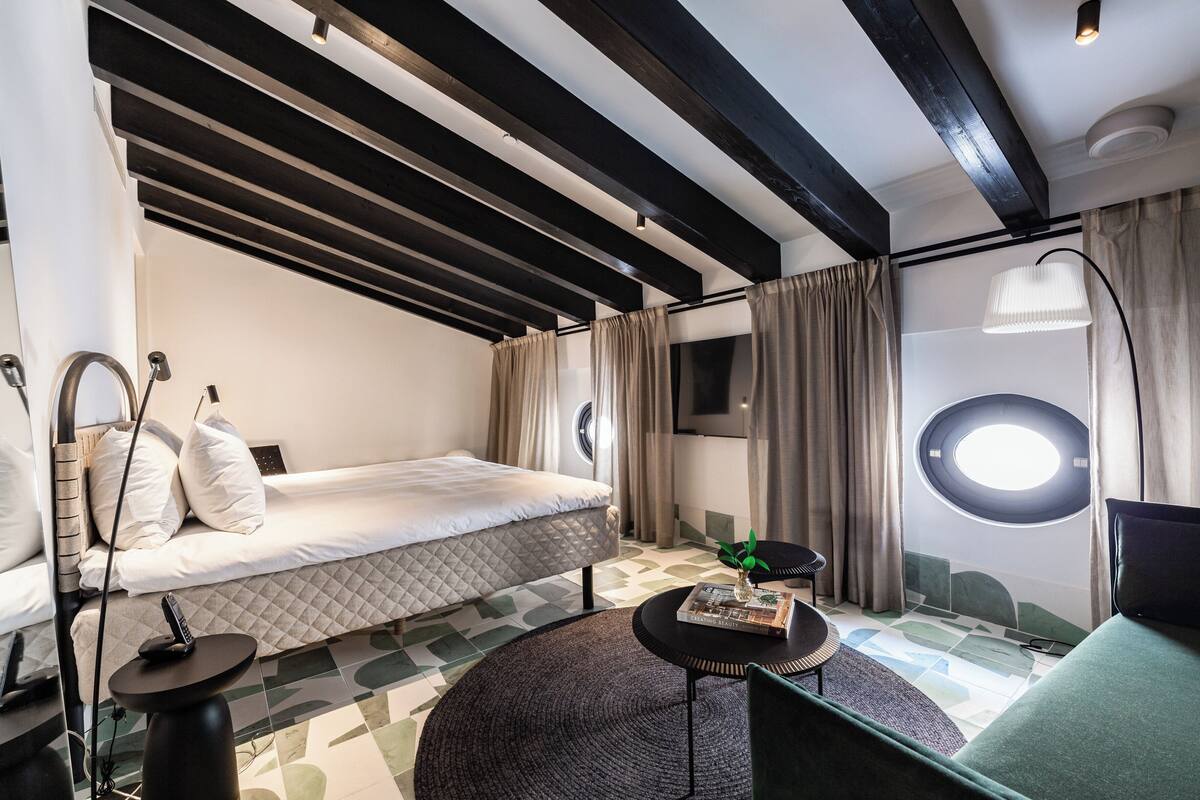 Ein modernes Schlafzimmer mit Bett, rundem Fenster, kleinem Tisch mit Pflanze und einem einzigartigen geometrischen Muster auf dem Boden.