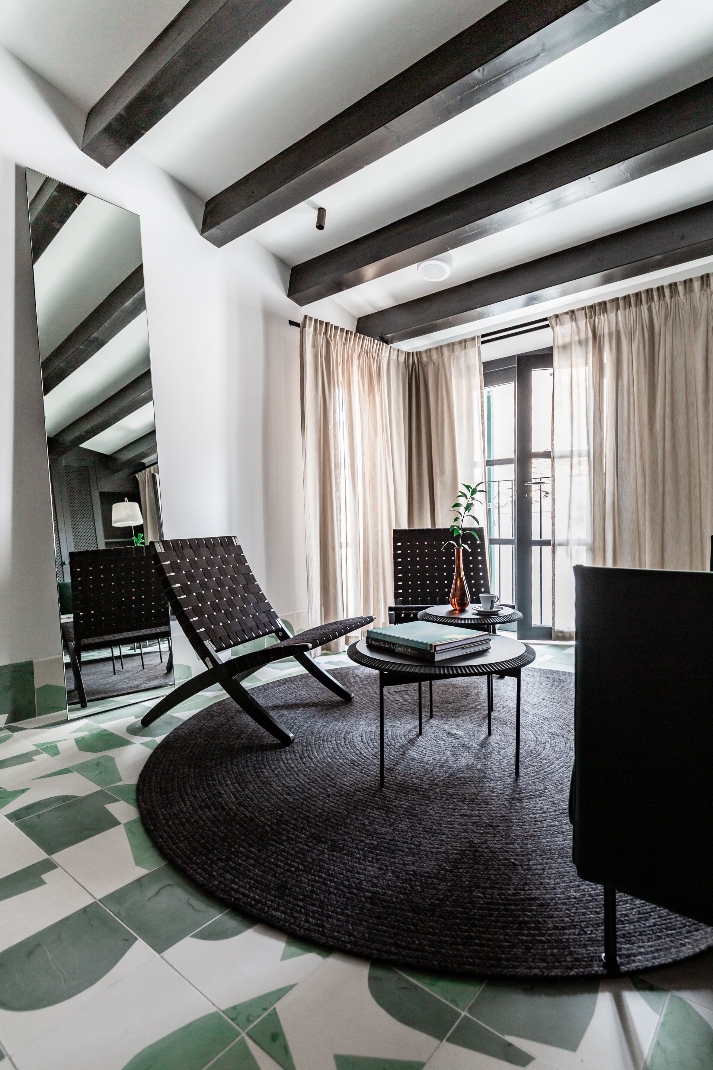 Une pièce moderne avec un plafond haut, des poutres apparentes, un tapis rond noir, deux fauteuils noirs, une petite table avec un vase et des livres, et une fenêtre avec des rideaux.