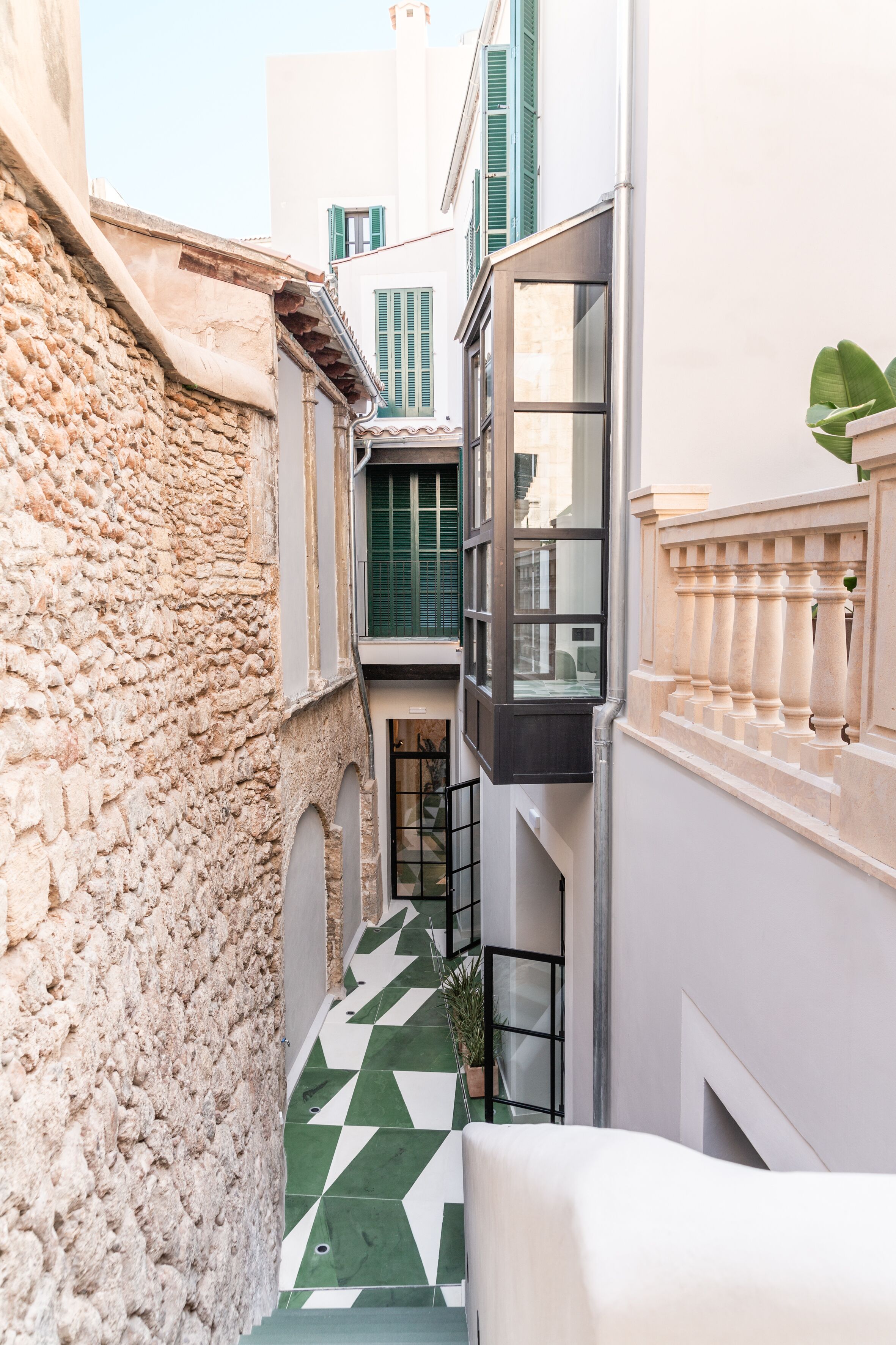 Un callejón estrecho con un muro de piedra a un lado y un edificio con piso de baldosas verdes y un balcón blanco al otro.
