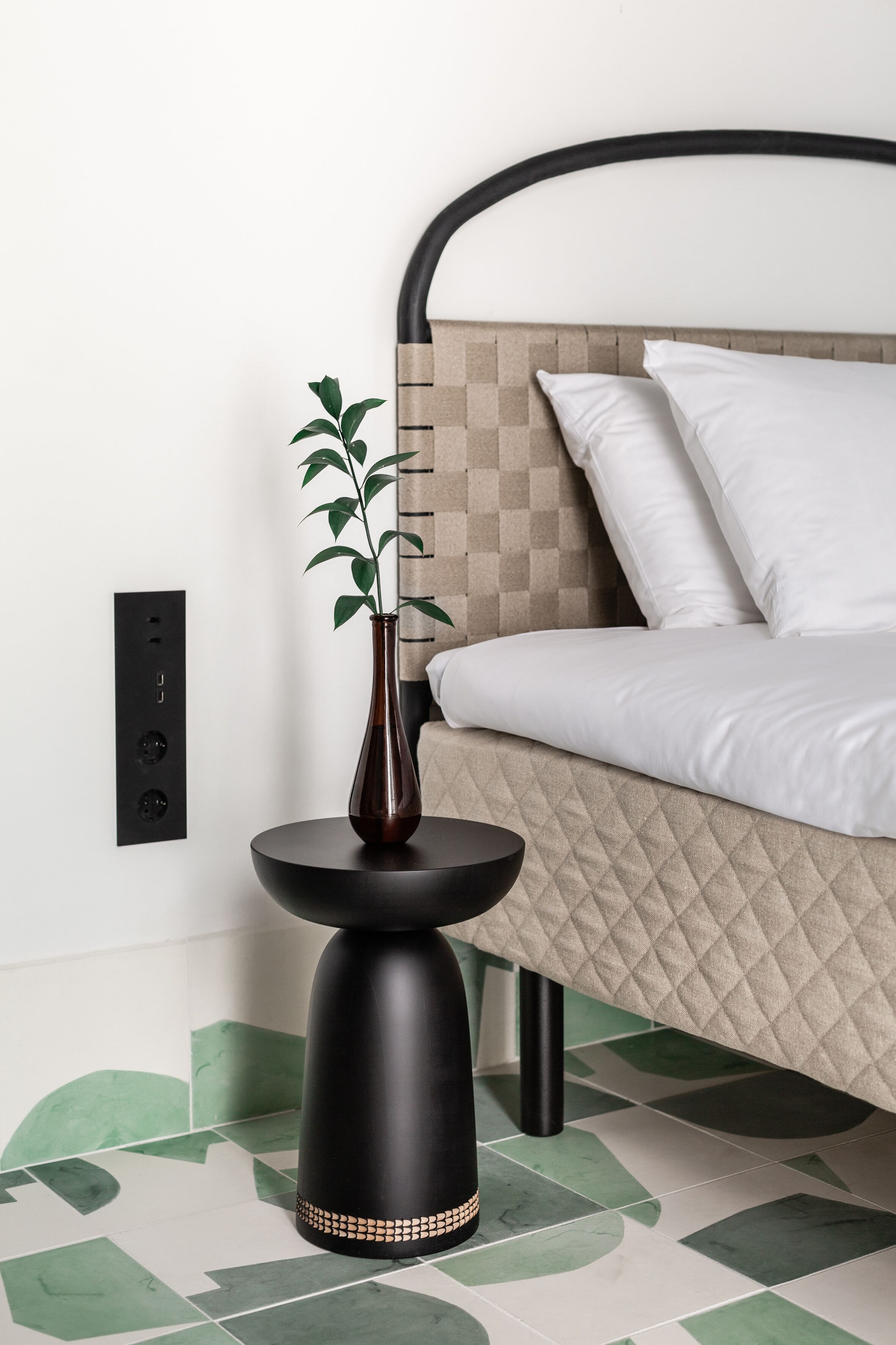 A modern bedroom with a bed, a bedside table with a vase, and a power outlet on the wall.