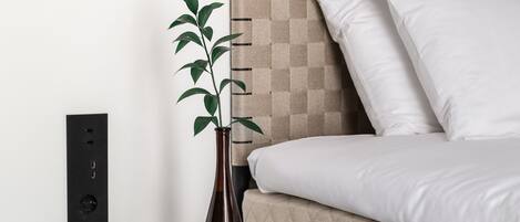 A modern bedroom with a bed, a bedside table with a vase, and a power outlet on the wall.