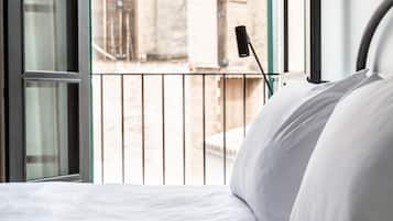 Una cama con ropa blanca, un ventanal y vista a un edificio de piedra con ventanas arqueadas.