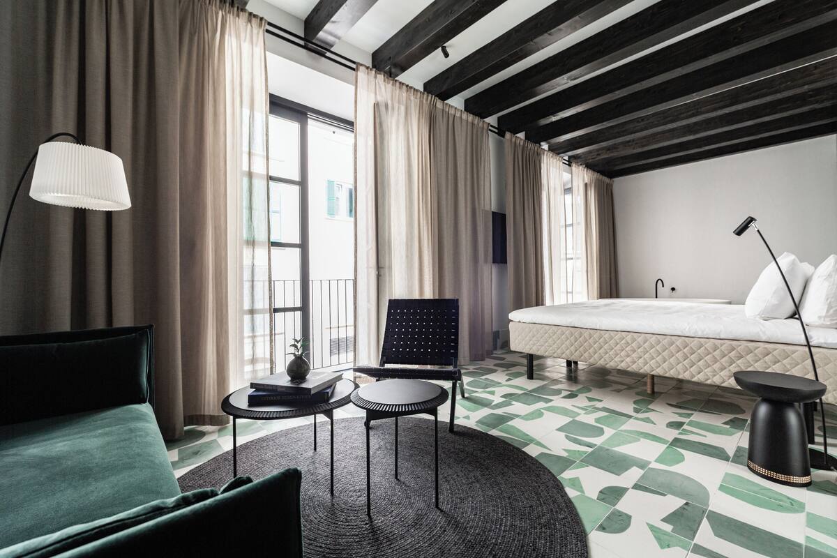 Ein modernes Hotelzimmer mit Bett, Sofa, rundem Tisch und Stuhl. Ein großes Fenster mit Vorhängen, ein Boden mit geometrischem Muster und eine Decke mit freiliegenden Balken.