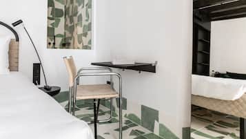 A hotel room with a bed, a desk with a chair, a mirror, a shelf with books, and a shower area.