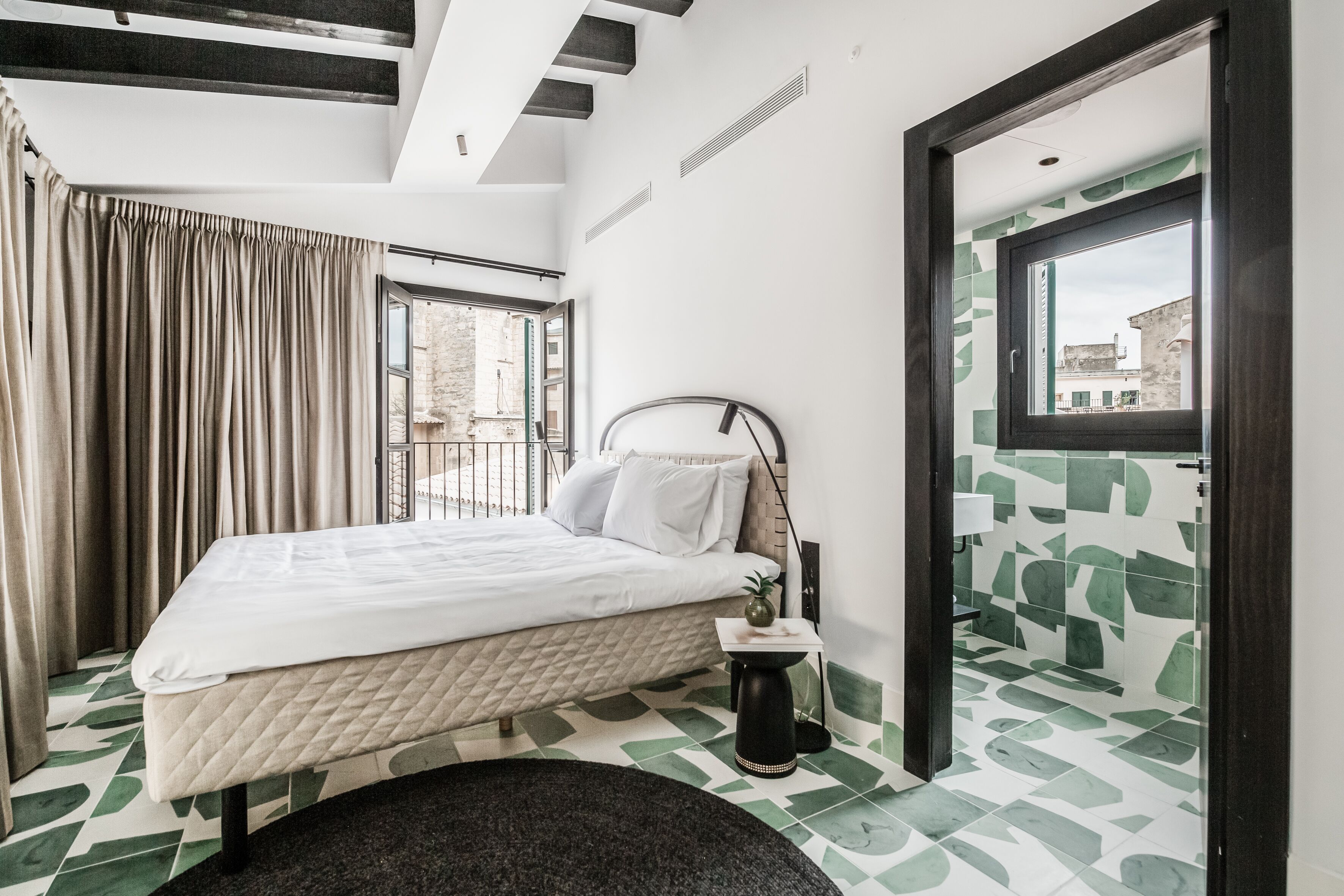 A bedroom with a bed, a bedside table, a window with curtains, and a view of buildings outside.