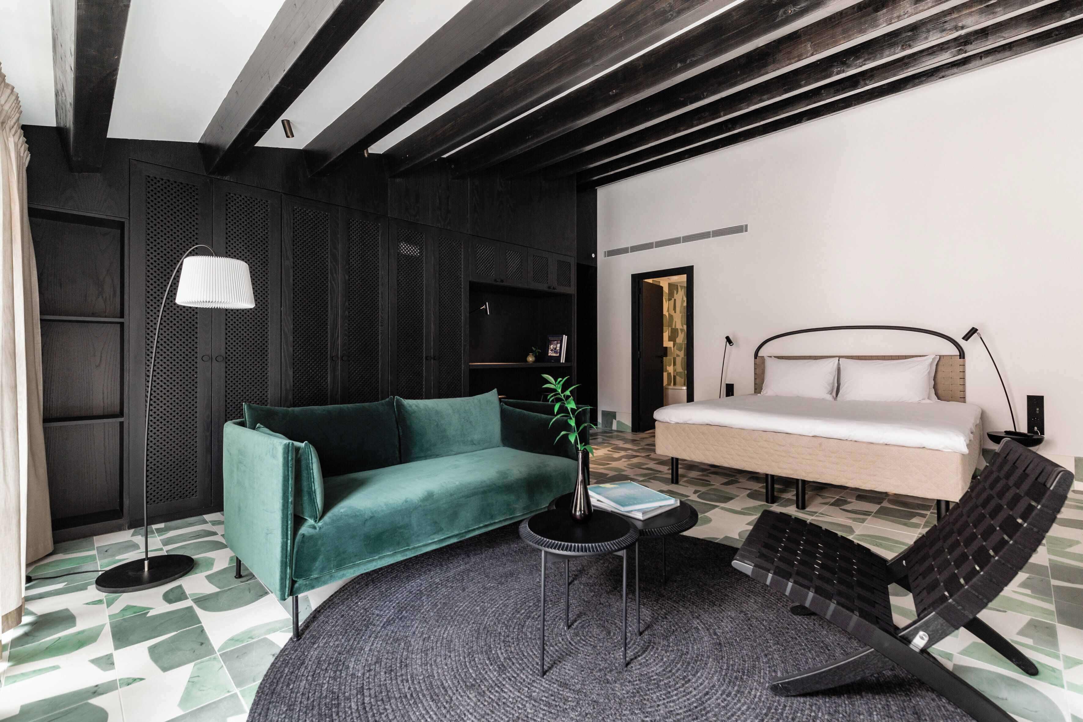 Ein modernes Zimmer mit einem grünen Sofa, einem kleinen runden Tisch mit einer Pflanze, einem schwarzen Sessel, einem Bett mit weißer Bettwäsche und einem gemusterten Boden.
