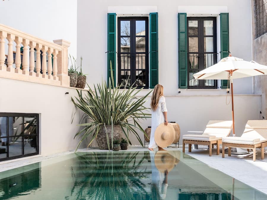 Una piscina con superficie de agua azul cristalina, una mujer con vestido blanco y un edificio blanco con contraventanas verdes.