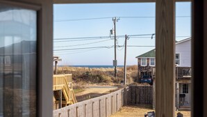 Blick durch ein Fenster auf eine Holzterrasse, einen Sandhof und ein Haus mit Balkon.