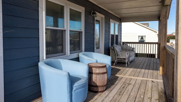 Eine überdachte Veranda mit blauen, gepolsterten Stühlen, einem Holzfass und einem Korbtisch.