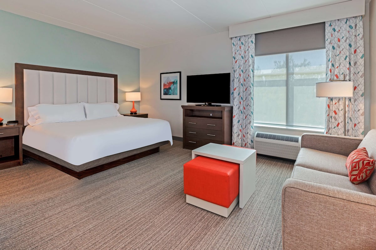 Ein Hotelzimmer mit einem großen Bett, einem Fernseher, einem Schreibtisch, einer Couch und einem Fenster mit Vorhängen.