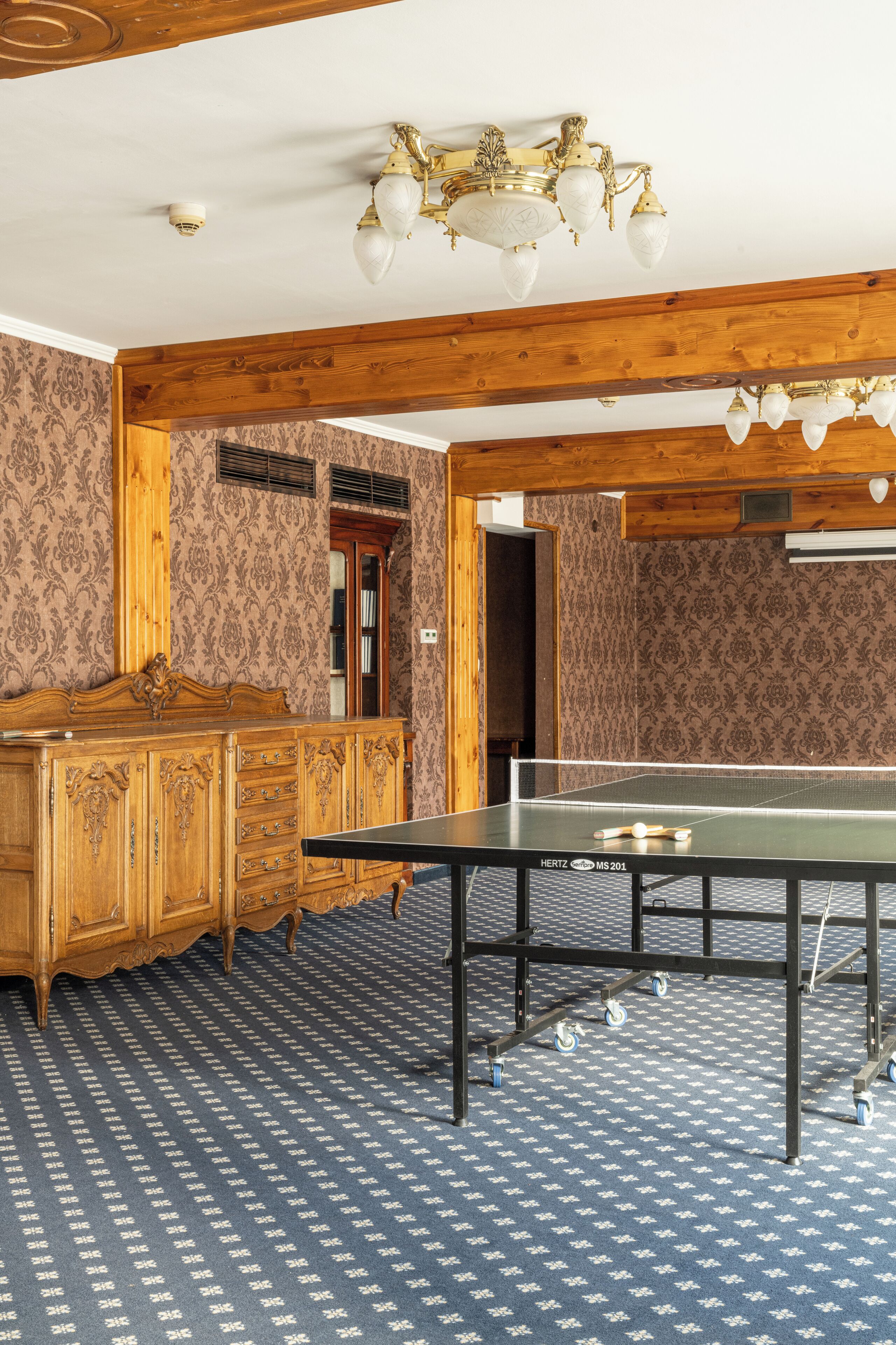 Ein Zimmer mit Tischtennisplatte, Holzmöbeln, gemustertem Wandpapier und einem Kronleuchter.
