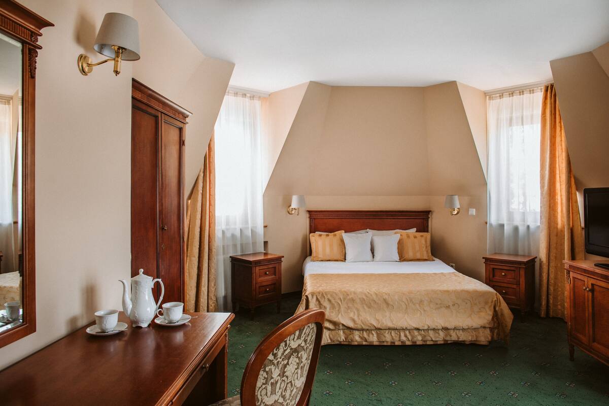 Una habitación de hotel con una cama, mesitas de noche, televisor, espejo y cómoda de madera.