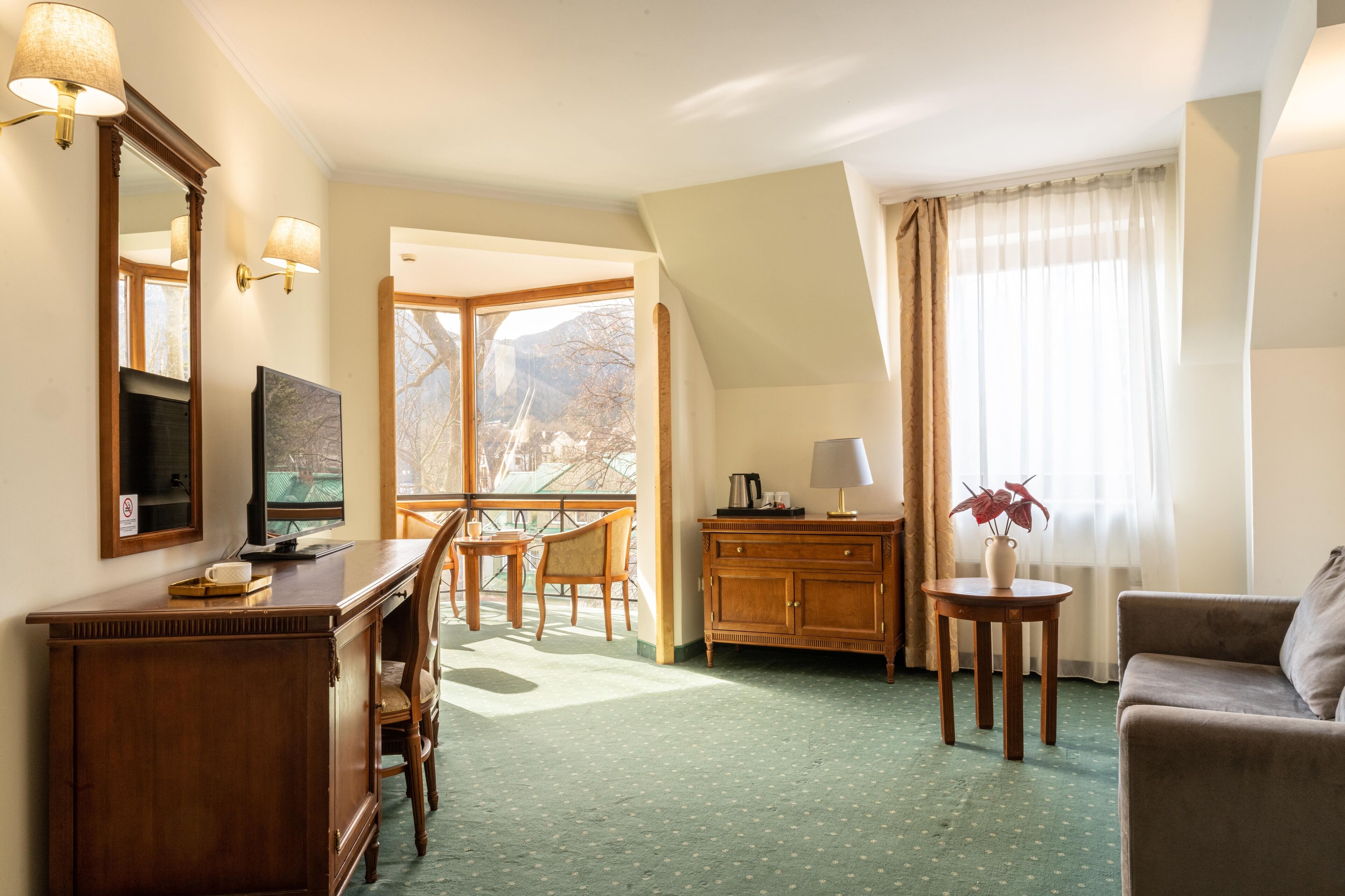 Een hotelkamer met een houten bureau, een televisie, een zithoek met stoelen, een bank, een kast, een bijzettafeltje met een vaas, en een raam met gordijnen.