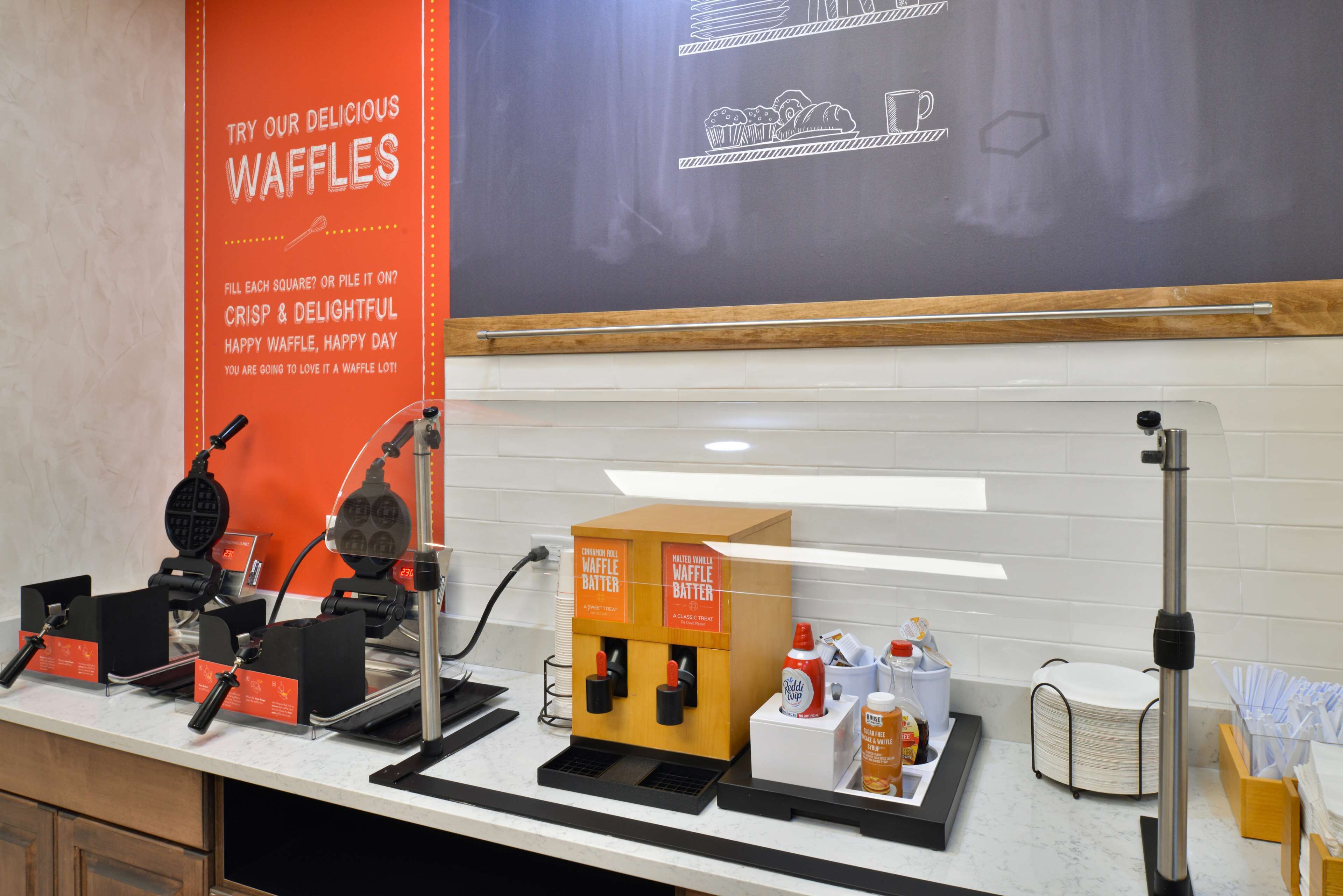 Una postazione per i waffle con due macchinette per waffle, un cartello pubblicitario che promuove i waffle e una lavagna con disegni a tema waffle.