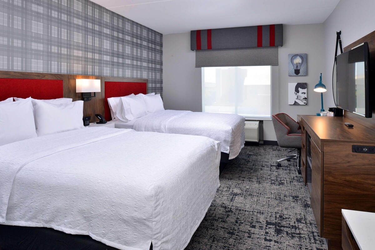 A hotel room with two beds, a desk, a chair, a lamp, and a large window.