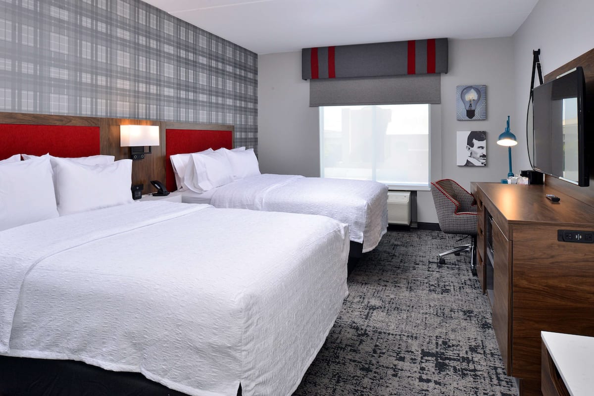 A hotel room with two beds, a desk, a chair, a lamp, and a large window.