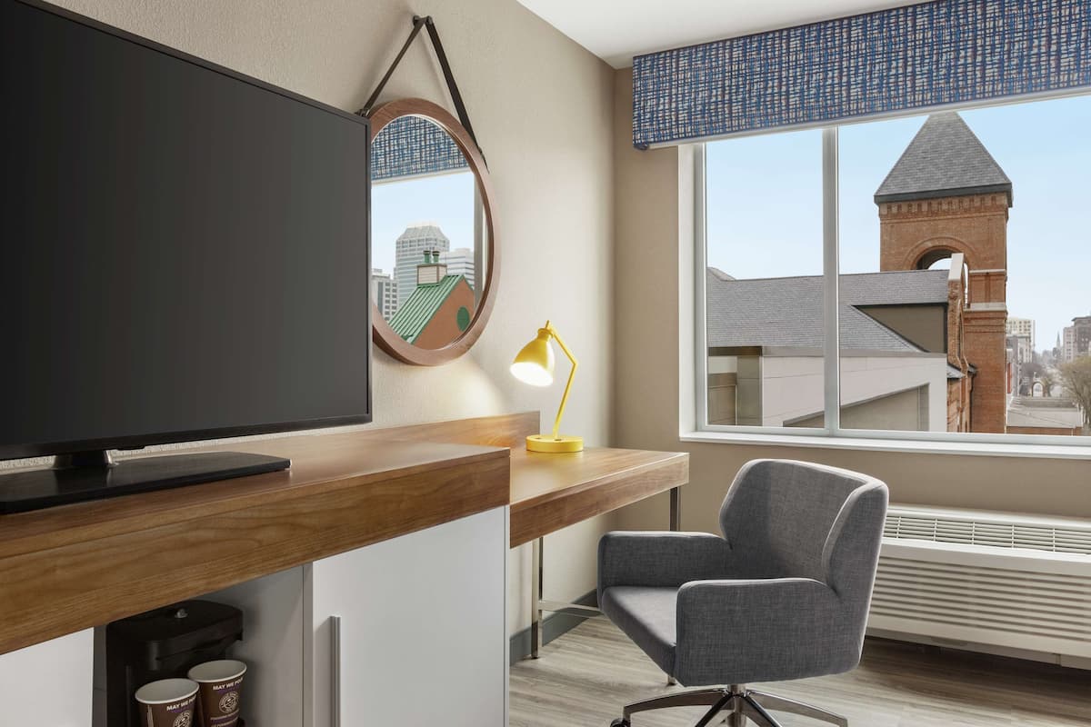 A modern hotel room with a large flat-screen TV, a wooden desk, a comfortable chair, a yellow desk lamp, and a view of a cityscape through a large window.