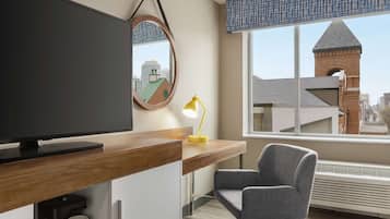 A modern hotel room with a large flat-screen TV, a wooden desk, a comfortable chair, a yellow desk lamp, and a view of a cityscape through a large window.