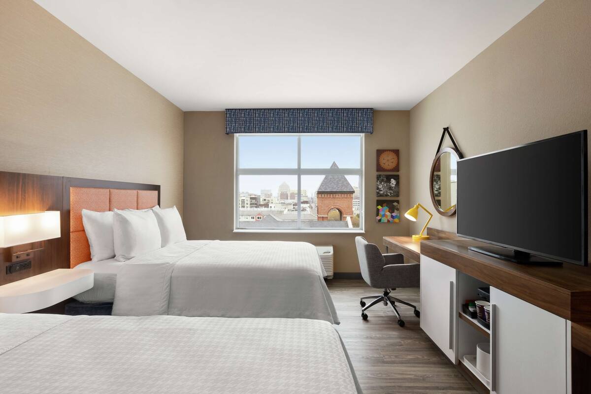 A hotel room with two beds, a desk with a TV, a chair, and a large window with a city view.