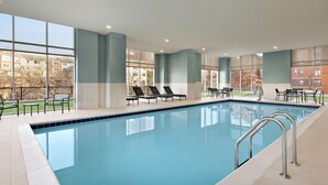 A swimming pool with lounge chairs and tables, large windows, and a view of buildings outside. - Hampton Inn Indianapolis Downtown IUPUI (Indianapolis)