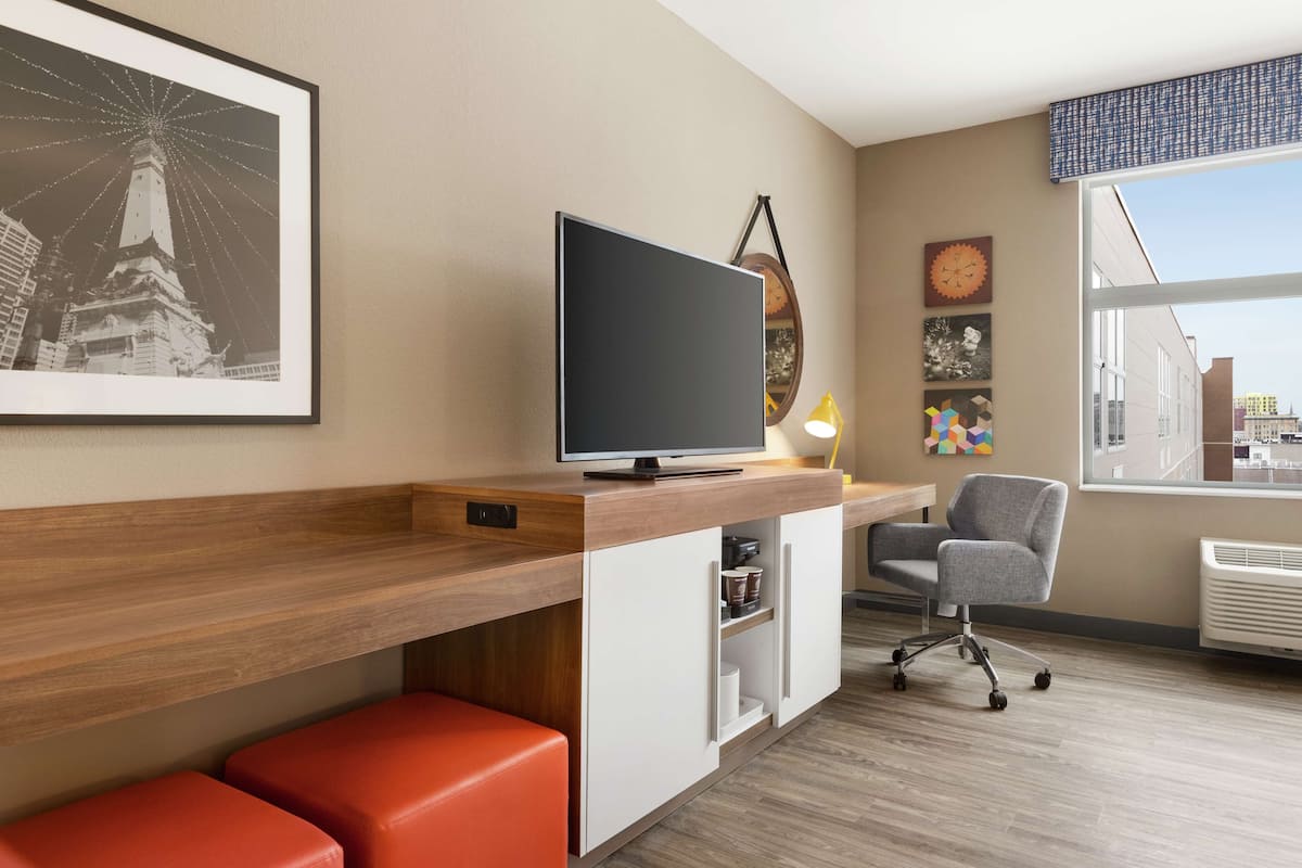 A modern hotel room with a wooden desk, a flat-screen TV, a comfortable chair, a red bench, and a large window offering a city view.