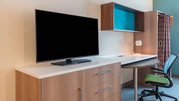 Une chambre d’hôtel moderne avec un téléviseur à écran plat posé sur un bureau blanc, une commode en bois avec des tiroirs, un fauteuil de bureau vert et un mur d’accent de couleur turquoise.