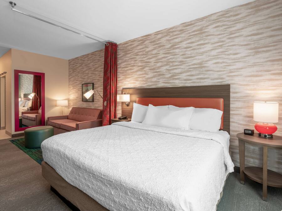 Et hotelværelse med en stor seng, et opholdsområde med en lænestol og skammel, et lille bord med lampe og et spejl med rød ramme.