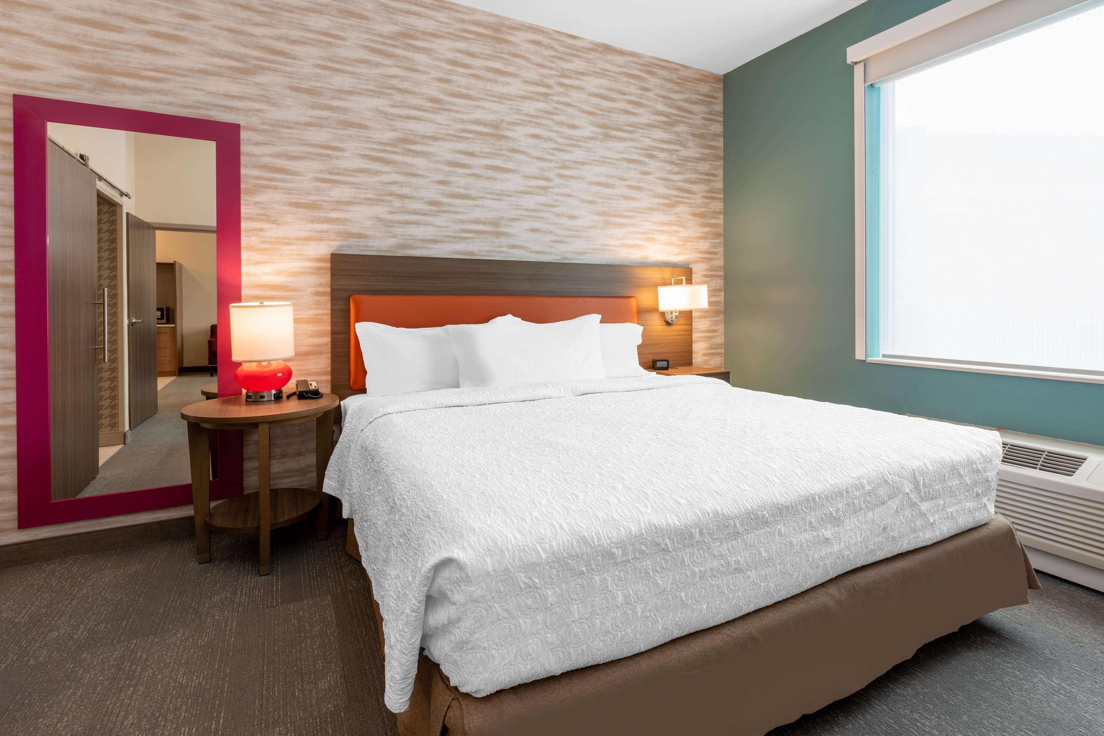 A hotel room with a large bed, a nightstand with a lamp, a mirror, and a window with a view.